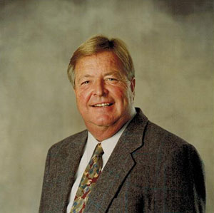 The image shows a man standing against a neutral background, wearing a suit with a tie, and smiling slightly at the camera.