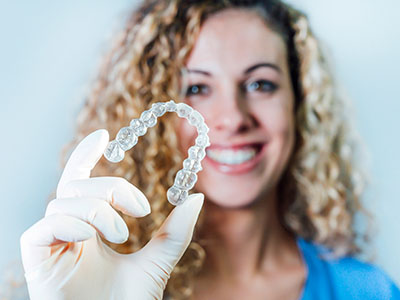 A woman wearing gloves holds up a large clear acrylic retainer with teeth-like shapes, smiling at the camera.