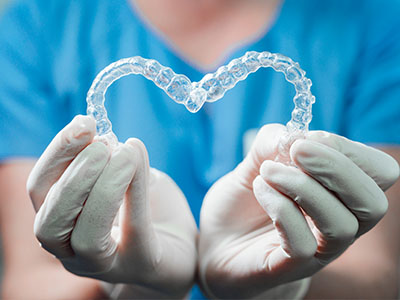 A hand holding a heart-shaped clear plastic object, which appears to be a dental appliance, against a blue background with a person wearing white gloves and a blue apron behind the hands.