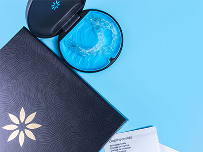 The image shows a blue box with an open lid containing a dental impression tray, a toothbrush, and a small container of crystals, all placed on a light blue surface against a dark background.
