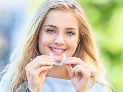 A young woman with blonde hair is smiling, holding a clear braces appliance up to her teeth while outdoors during daytime.