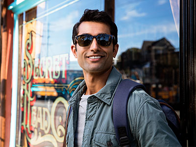A man wearing sunglasses and a backpack stands outside a storefront, smiling towards the camera.