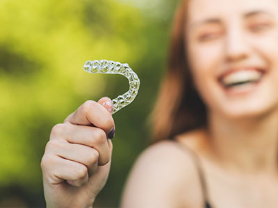 An image featuring a young woman holding up a clear plastic toothbrush with her left hand, smiling broadly, against a blurred outdoor background.