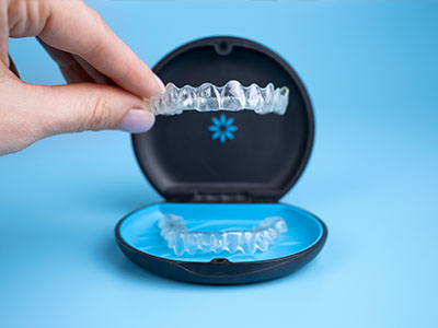 A hand holding a clear plastic tray with a blue lid containing a full set of transparent dental implants.