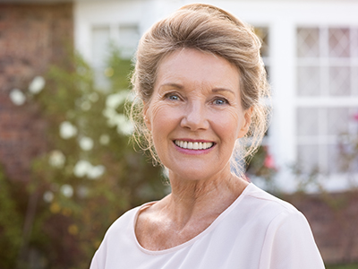 An elderly woman with blond hair wearing a pink top smiles at the camera while standing in front of a brick house with white trim.