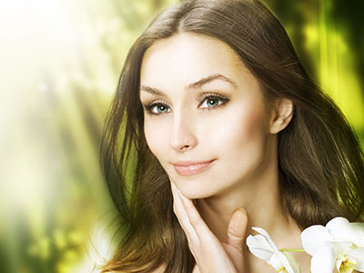 A woman with fair skin and blue eyes wearing makeup, with her hand gently resting on her chin, looking directly at the camera.