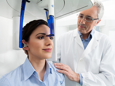 The image shows a woman seated in a dental chair with a blue device on her head, receiving care from a dentist who is standing nearby.