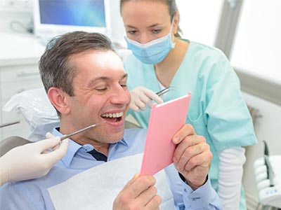 This is a photograph showing a man seated in a dental chair with a smile, looking at a pink card held by a woman who appears to be a dental professional. The setting suggests a dental office environment, indicated by the presence of medical equipment and the attire of the individuals present.