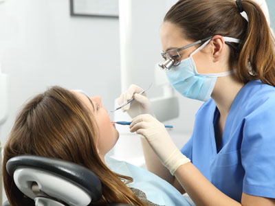 A dental hygienist working on a patient s teeth with dental tools, wearing personal protective equipment and a blue surgical gown, in a dental office setting.