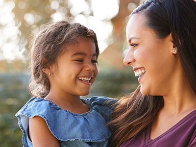 A woman and a young child are smiling at each other outdoors during daylight.