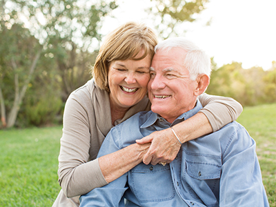 The image shows an older couple embracing each other outdoors, with the man wearing glasses and the woman with short hair. They appear to be enjoying a sunny day together.