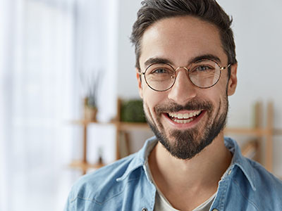 The image shows a man with a beard smiling at the camera, wearing glasses, a blue shirt, and a light-colored denim jacket, standing against a blurred background that suggests an indoor setting.