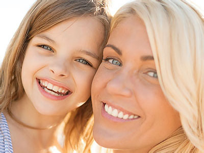 The image shows two individuals, an adult woman with light skin and blue eyes, smiling at the camera, with a young child beside her, both sharing a joyful moment outdoors.