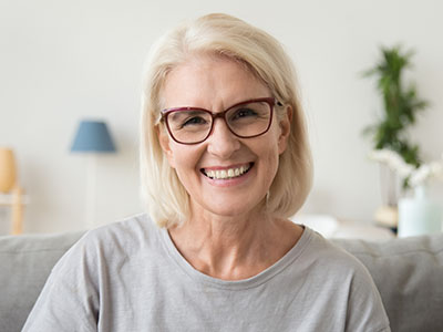 A smiling woman with short blonde hair, wearing glasses and a light-colored top, seated on a couch indoors.