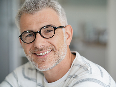 The image features a man with grey hair, wearing glasses, smiling at the camera with his head tilted slightly to the side. He has a beard and mustache, and he s dressed in a casual white t-shirt.