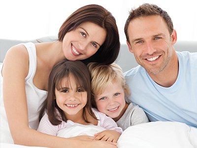 A family of four posing together on a bed with smiles and relaxed expressions.