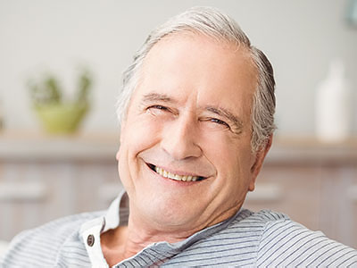 A smiling older man with gray hair, wearing a blue shirt, sitting in a comfortable chair indoors.