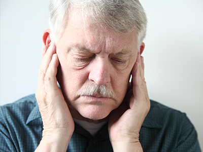 The image shows an elderly man with his eyes closed, holding his face with both hands near his ears, appearing to be in a state of distress or discomfort.