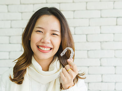 The image shows a smiling woman holding up a dental retainer with her right hand. She has dark hair, wears a light-colored top, and stands against a brick wall background.